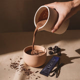 Hot chocolate poured from a jug into a cup, with a Serune sachet, cherries and vanilla alongside