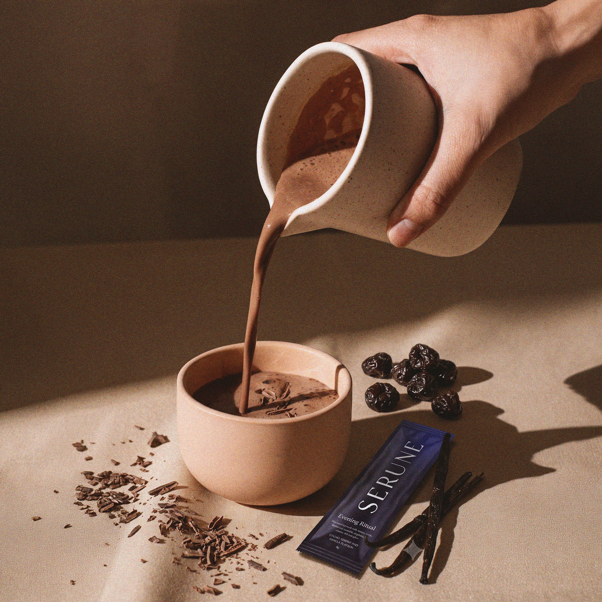 Hot chocolate poured from a jug into a cup, with a Serune sachet, cherries and vanilla alongside