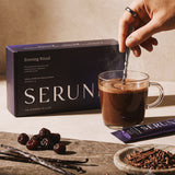 Stirring a glass mug of Serune hot chocolate in front of the box, with cherries and vanilla on the table